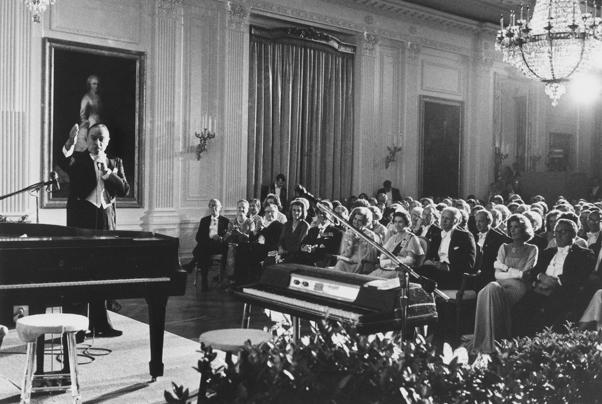 This photograph depicts President Gerald Ford and guests in the center and right of image. A piano sits in the left foreground. Comedian Bob Hope is standing behind and left of piano, gesturing and speaking into microphone, during his monologue for the entertainment portion of the State Dinner held in honor of the Queen of England
