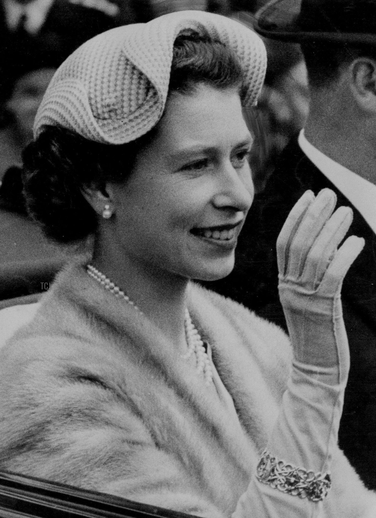 The Queen, smiling and wearing an attractive white wavy-edged hat, drives along the Ascot course with the Duke of Edinburgh on this final day of the Royal meeting. June 18, 1954